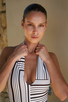 A woman wearing a black and white vertically striped halter-neck swimsuit, posing with her hands on the straps against a neutral beige background.