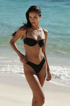 A woman modeling a black two-piece bikini on a sandy beach with turquoise ocean water in the background.