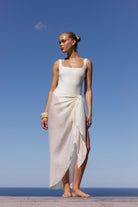 A woman stands barefoot outdoors against a clear blue sky and ocean, wearing a sleeveless white wrap-style dress with a square neckline and draped side detail.