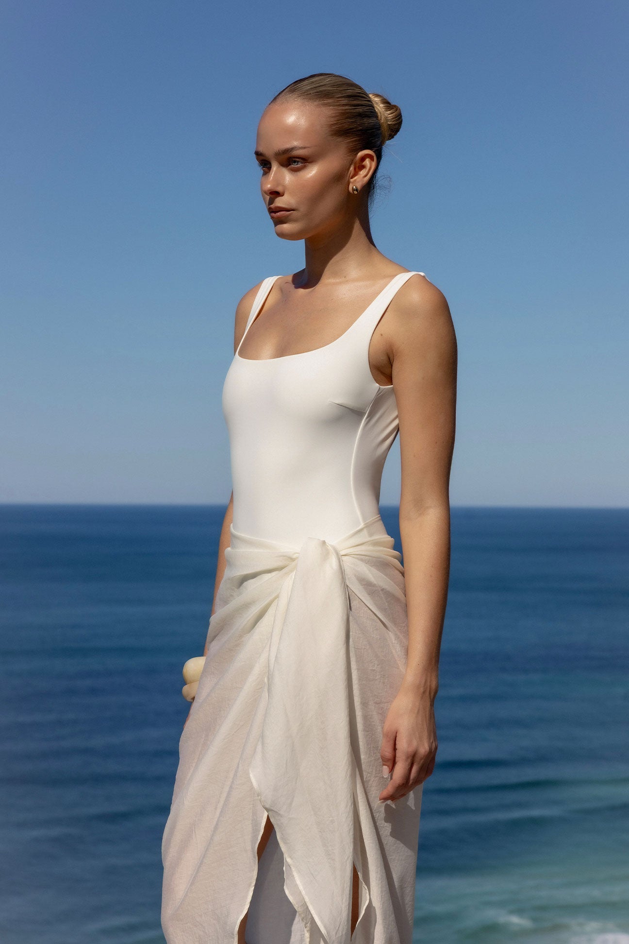 A woman stands outdoors by the ocean wearing a fitted white sleeveless bodysuit with a draped white wrap skirt tied at the waist. The background shows blue sky and sea.