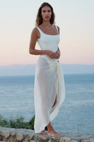 A woman stands barefoot on a stone wall overlooking the sea at dusk, wearing a white sleeveless maxi dress with a side slit and a draped wrap skirt tied at the hip.