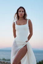 A woman wearing a white sleeveless dress with a square neckline and a draped side slit, standing outdoors against a soft sky and distant water background.