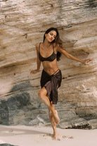 Woman in a brown bikini and sarong standing against a stone wall.