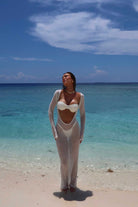 Woman in a white outfit standing on a beach with clear blue water and sky.