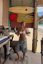 Woman in a bikini top and shorts standing on a sandy beach with surfboards stacked behind her.