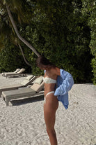 Person in a bikini standing on a sandy path with lounge chairs and greenery in the background
