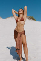 Woman in a brown bikini and sarong standing on a sandy beach with a clear blue sky.