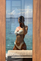 Woman in a white bikini standing by a large window with a view of the ocean.