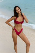 Woman in a magenta bikini standing on a beach with ocean waves in the background
