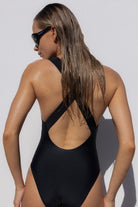 A woman wearing a black one-piece swimsuit, photographed from the back against a light gray wall. The swimsuit has a large open back with crisscrossing straps.