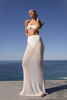 A woman stands outdoors by the ocean wearing a white bandeau bikini top and a long sheer white cover-up skirt. The background shows a clear blue sky and sea, with a concrete ledge beneath her.